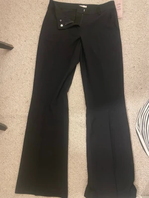 Svarta bootcut kostymbyxor Gina Tricot - Snygga svarta kostymbyxor från Gina Tricot med bootcut passform och hög midja. Byxorna har bälteshällor, två bakfickor och stängs med knapp och dragkedja. Perfekta för en clean och stilren look.