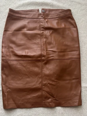 Brun pennkjol i skinnimitation - Snygg brun pennkjol i skinnimitation med hög midja och slits bak. Kjolen har dragkedja och knapp bak för enkel påklädning. Perfekt för en trendig och stilren look.