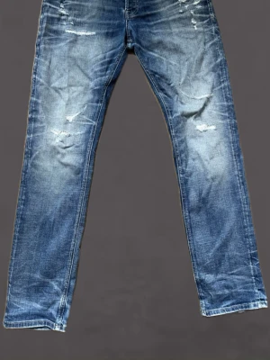 Blå slitna raka jeans - Säljer ett par blå jeans med raka ben och slitna detaljer framtill. Jeansen har en klassisk femficksmodell och är tillverkade i denim med snygg tvätt och slitningar för en avslappnad look.