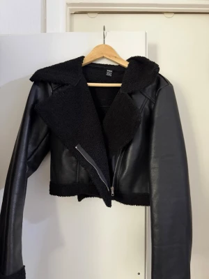 Svart kort fuskpäls-cardigan H&M - Snygg svart cardiganjacka från H&M i kort modell med fuskpälsdetaljer på krage, insida och nederkant. Jackan har dragkedja framtill och är gjord i ett läderliknande material som ger en cool look. Perfekt för dig som vill ha en trendig och varm jacka.