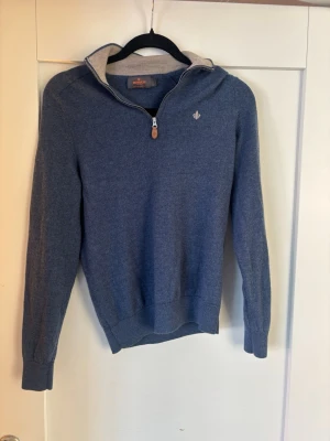 Blå halvzip tröja från Morris - Snygg blå stickad tröja från Morris med halv dragkedja och kontrastfärgad insida på kragen. Tillverkad i 100% merinoull och har en liten broderad logga på bröstet. Perfekt för lager på lager och riktigt skön att bära.