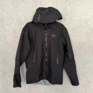 Arc'teryx Beta LT Jacket - Snygg svart vindjacka från Arc'teryx med huva och dragkedjor framtill. Jackan har två stora fickor med dragkedja och en diskret logga på bröstet. Tillverkad i ett lätt och slitstarkt material som står emot vind och regn. Perfekt för utomhusaktiviteter.