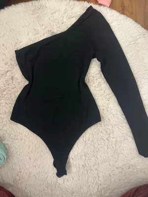 Svart one shoulder body - Snygg svart body med one shoulder-design och lång ärm på ena sidan. Tillverkad i stretchigt material som sitter tight och framhäver figuren. Perfekt för att skapa en trendig och stilren look.
