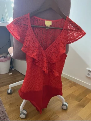 Röd spetsbody med volang från H&M - Snygg röd body från H&M i spets med volangdetaljer framtill och vid ärmarna. Djup V-ringning och korta, fladdriga ärmar. Tillverkad i genombruten spets och stretchigt tyg nedtill för bekväm passform. Perfekt för att lyfta vilken outfit som helst.