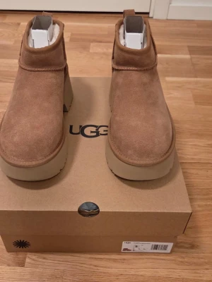 Bruna platåstövletter från UGG - Säljer ett par snygga bruna stövletter från UGG med hög platåsula och rund tå. Skorna är i mocka med mjukt foder och har en chunky siluett. Perfekta för dig som vill ha både stil och komfort. Klassisk UGG-logga bak på hälen.