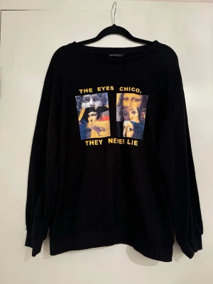 Svart sweatshirt med tryck - Cool svart hoodie med färgstarkt tryck på bröstet och texten 'The eyes chico, they never lie' i gult. Tröjan har rund halsringning och ribbade muddar. Materialet är mjukt och bekvämt, perfekt för att ge din outfit en unik touch.