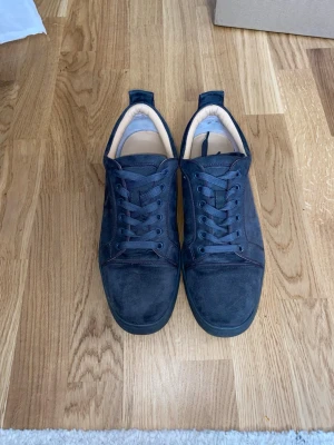 Grå louboutin  - Snygga gråa sneakers i mocka med klassisk snörning och låg profil. Skorna har en unik röd yttersula som sticker ut och ger en cool kontrast. Insidan är ljus och mjukt vadderad för extra komfort. Perfekta för dig som vill ha stilrena men ändå unika sneakers.