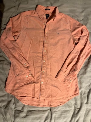 Rosa gant skjorta  - Säljer en laxrosa skjorta från Gant med klassisk krage och knappar framtill. Skjortan har lång ärm och liten broderad logga på bröstet. Tillverkad i mjuk bomull och har en avslappnad passform som funkar till många olika stilar.