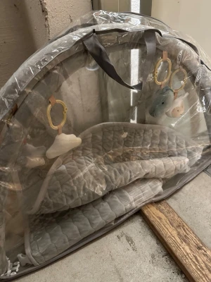 Grå babygym med leksaker - Säljer ett grått babygym med quiltad matta och mjuka hängande leksaker i form av moln och djur. Babygymmet har en båge och kommer i en genomskinlig förvaringsväska med handtag. Perfekt för lek och stimulans för små barn.