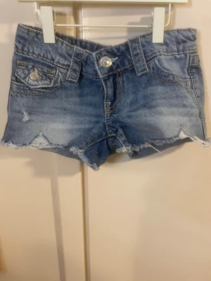 Blå jeansshorts med rå kant - Har använt 1-2 gånger. Snygga blå jeansshorts med fransig, rå kant och slitningar framtill. Klassisk femficksmodell med silverfärgade knappar och bälteshällor. Perfekta för varma dagar och enkel att matcha med olika toppar.