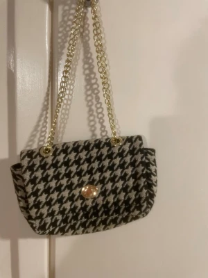 Houndstooth handväska från H&M - Snygg handväska från H&M i klassiskt svartvitt hundtandsmönster. Väskan har guldfärgad kedjerem och ett elegant guldfärgat spänne framtill. Materialet är textil med struktur och väskan har en praktisk innerficka. Perfekt accessoar för att lyfta din outfit.