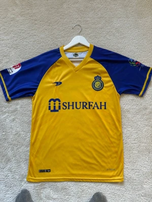 Al Nassr matchtröja Ronaldo #7 - Snygg gul och blå Al Nassr matchtröja med Ronaldo och nummer 7 på ryggen. Tröjan har klubbmärke och sponsorlogga på bröstet, samt tryck på ärmarna. Tillverkad i lätt och ventilerande material, perfekt för fotbollsträning eller match.