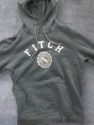Grå hoodie från Abercrombie & Fitch - Mysig grå hoodie från Abercrombie & Fitch med stor magficka, dragsko i huvan och broderad text 'FITCH' samt emblem på bröstet. Perfekt för chill dagar och streetwear-stil. Tillverkad i mjukt bomullsmaterial.