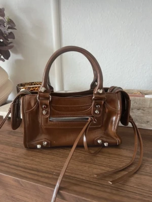 Brun minihandväska i läder - Snygg brun minihandväska i glansigt läder med silverfärgade detaljer och dragkedja framtill. Väskan har dubbla handtag, dekorativa nitar och långa läderband. Perfekt för dig som gillar trendiga accessoarer med edgy känsla.