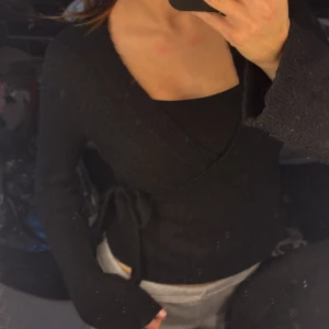 Svart omlottopp med knytband - Snygg svart omlottopp med långa ärmar och knytband i midjan. Tröjan har en djupare v-ringning och är gjord i ett mjukt, stickat material. Toppen är lite nopprig men inget ja skulle tänka på.