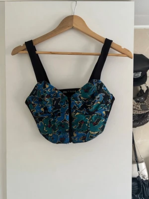 Blommig korsettopp från Zara - Snygg korsettopp från Zara med breda axelband och blommigt mönster i blått, grönt och svart. Toppen har en croppad passform och coola detaljer framtill. Perfekt att styla med ett par snygga jeans eller kjol för en trendig look. 