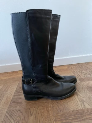Svarta läderstövlar  - Snygga svarta stövlar i läder med stretchigt tyg längs skaftet och dekoration vid ankeln. Klassisk damstövel med låg klack och dragkedja på insidan. En stilren stövel. Sköna att gå i. Använda men i bra skick. Stl 39. 