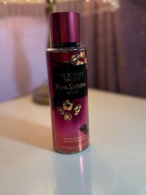 Victoria's Secret Pure Seduction Noir - En lyxig doftmist från Victoria's Secret i serien Pure Seduction Noir. Flaskan är mörklila med guldiga blommor och svart etikett. Innehåller 250 ml och har en elegant, feminin design som passar perfekt på sminkbordet.