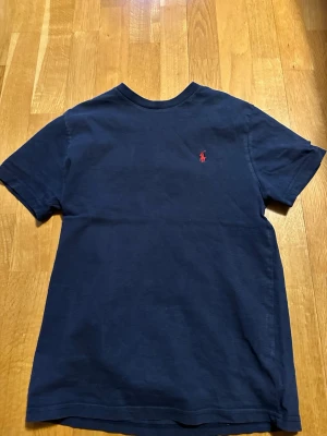 Marinblå t-shirt från Polo Ralph Lauren - Klassisk marinblå t-shirt från Polo Ralph Lauren med röd broderad logga på bröstet. T-shirten har rund halsringning och korta ärmar. Tillverkad i mjuk bomull för en skön känsla och enkel stil.