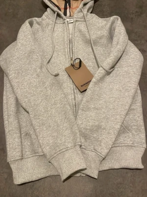 Ljusgrå zip hoodie från Burberry  - Snygg ljusgrå hoodie från Burberry med dragkedja framtill och klassisk huva med snören. Hoodien har ribbade muddar vid ärmar och nederkant samt fickor på sidorna. Insidan av huvan har Burberrys ikoniska rutmönster. Tillverkad i mjukt bomullsmaterial.