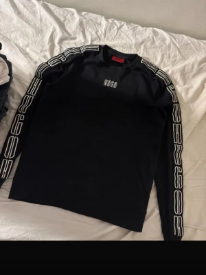 Svart sweatshirt från Hugo - Säljer en svart sweatshirt från Hugo med vita logotyper längs ärmarna och tryckt Hugo-text på bröstet. Tröjan har rund halsringning och ribbade muddar. Perfekt för dig som gillar streetwear och enkel, stilren design.