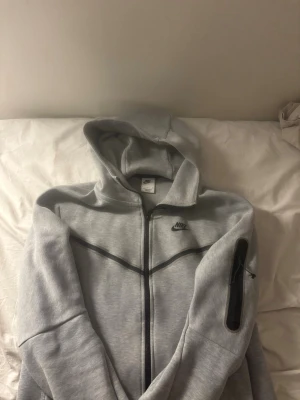 NIKE TECH hoodie, ordinarie pris 1099kr - Snygg grå hoodie från Nike med hel dragkedja och svart logga på bröstet. Hoodien har svarta detaljer längs bröstet och en praktisk ficka med dragkedja på ärmen. Tillverkad i mjukt material och har en klassisk huva.