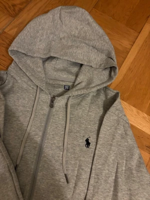 Grå loungewear-set från Polo Ralph Lauren - Mysigt grått loungewear-set från Polo Ralph Lauren med dragkedja på hoodien och matchande byxor med dragsko. Båda delarna har den klassiska Polo-loggan broderad på bröstet och benet. Tillverkad i mjuk bomullsblandning, perfekt för chill dagar hemma.