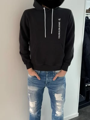 Svart Calvin Klein Jeans hoodie - Snygg svart hoodie från Calvin Klein Jeans med vit logga tryckt vertikalt både fram och bak. Hoodien har huva med vita dragsnören och en stor magficka. Tillverkad i mjukt bomullsmaterial som är skönt att bära. Perfekt för en avslappnad och trendig look. Storlek S men passar mig som har m, lite urtvättad.