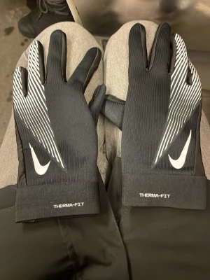 Nike Therma-Fit svarta handskar - Snygga svarta Nike Therma-Fit handskar med vitt mönster och logga på ovansidan. Handskarna har greppvänlig insida och är perfekta för kyliga dagar. De har en sportig look med vita detaljer och ribbade muddar.