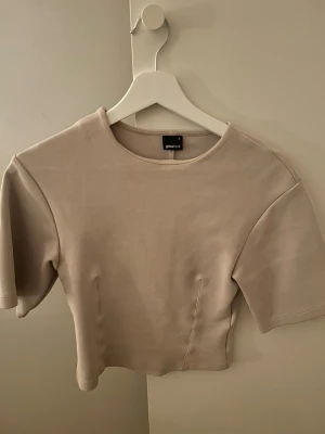 Beige croppad topp från Gina Tricot - Snygg beige croppad topp från Gina Tricot med rund halsringning och korta ärmar. Toppen har en stilren och enkel design som passar till mycket. Materialet känns mjukt och stretchigt, perfekt för en trendig vardagslook.