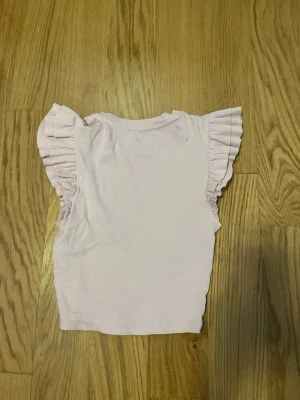 Ljusrosa ribbad topp med volangärm - Supersöt ljusrosa topp från Zara med ribbad struktur och volangdetaljer vid ärmarna. Toppen är ärmlös men har breda volangärmar som ger en extra touch. Perfekt för dig som gillar en feminin och trendig stil.