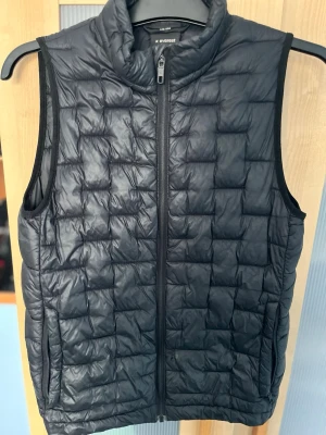 Svart dunväst från Everest - Stilren svart dunväst från Everest med quiltat rutmönster och hög krage. Västen har dragkedja framtill och två sidofickor. Perfekt att slänga över en hoodie eller tröja för extra värme. Materialet är lätt och smidigt, vilket gör den enkel att bära med sig.