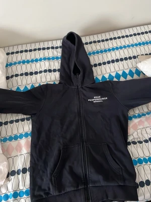 Svart Peak Performance hoodie - Säljer en svart hoodie från Peak Performance med dragkedja och fickor framtill. Märket syns tryckt i vitt på bröstet. Klassisk modell med huva och ribbade muddar. Perfekt för chill dagar eller när du vill ha en enkel och snygg look. passar 160cm