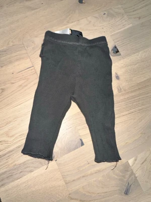 Mörkgrå ribbade leggings med volang - Säljer ett par mörkgrå leggings i ribbat material med söt volangdetalj bak vid midjan. De har stretchig midja och råa kanter vid bensluten för en avslappnad look. Perfekta att matcha med oversized tröjor eller hoodies.