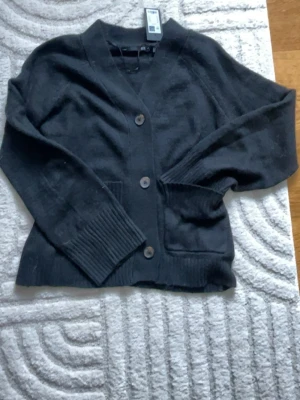 Svart kofta med knappar - Säljer en svart kofta med v-ringning och stora knappar framtill. Koftan har två fickor och breda ribbade muddar vid ärmslut och nederkant. Perfekt att slänga över en t-shirt eller topp för en chill look. Helt oanvänd har kvar prislappen