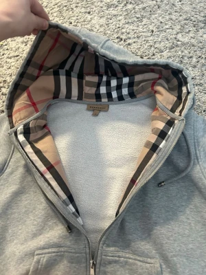Grå zip hoodie från Burberry - Snygg grå hoodie med dragkedja från Burberry. Klassisk huva med det ikoniska rutmönstret på insidan, dragsko och två stora fickor framtill. Tillverkad i mjukt sweatshirtmaterial, perfekt för en chill och stilren look.