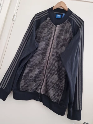 Adidas Originals träningsjacka 2XL - Snygg Adidas Originals träningsjacka i mörkgrå/svart med unikt mönstrat framparti. Klassiska tre ränder längs ärmarna och dragkedja hela vägen framtill. Storlek 2XL. Säljs pga stor viktnedgång.  I riktigt fint skick – inga fläckar eller skador. Enda lilla anmärkningen är att det grå partiet runt kragen har börjat flagna/ljusna lite, men det syns inte vid användning.   Detta är ett plagg som är riktigt svårt att få tag på idag – perfekt för samlare och adidas-fans