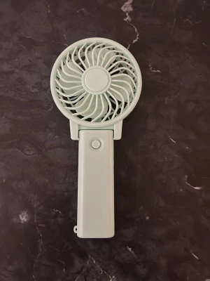 Mintgrön portabel handfläkt - Smidig mintgrön portabel handfläkt i plast med vikbart handtag. Fläkten har en enkel knapp för att slå på och av, och kan både hållas i handen eller ställas på ett bord. Perfekt när du behöver lite extra svalka på språng eller vid skrivbordet.