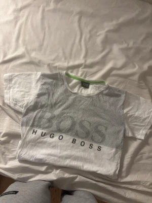 Vit mesh t-shirt från Hugo Boss - Säljer en vit t-shirt från Hugo Boss med meshdetaljer över bröstet och stort BOSS-tryck. Under meshpartiet står HUGO BOSS tryckt i svart. T-shirten har rund halsringning med grön insida och korta ärmar. Perfekt för en clean och sportig look.