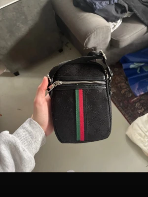 Svart axelväska med röd/grön rand - Snygg svart axelväska från Gucci med präglat GG-mönster och en ikonisk röd och grön rand framtill. Väskan har en justerbar axelrem och en silverfärgad dragkedja. Perfekt storlek för det viktigaste och ger en lyxig touch till din outfit.