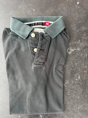 Svart piké t-shirt med krage - Klassisk svart piké t-shirt med korta ärmar och två knappar framtill. T-shirten har en stilren krage i matchande färg och är tillverkad i ett mjukt bomullsmaterial. Perfekt för en avslappnad men ändå snygg look.