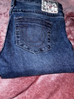 Blå True Religion jeans med brodyr - Säljer ett par klassiska blå jeans från True Religion med ikonisk broderad bakficka och tydlig logga på etiketten. Jeansen har raka ben och är gjorda i slitstark denim med snygga kontrastsömmar. Perfekta för dig som gillar streetwear och vill sticka ut. VIKTIGT ATT VETA, byxorna är uppsydda i längden, de passar mig i längden som är ca 160cm. 