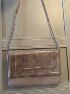 Rosa kuvertväska med axelrem - Säljer en snygg rosa kuvertväska med axelrem. Väskan har en skimrande yta med ormmönstrad struktur på locket och remmen, medan resten är i mjuk mockaliknande material. Perfekt för dig som vill ha en stilren och trendig accessoar.