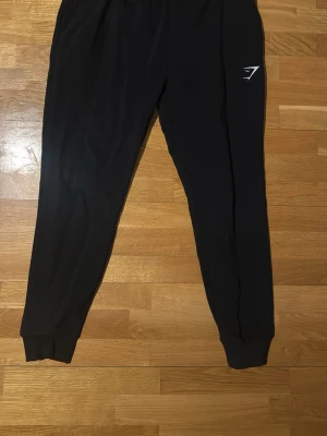 Svarta mjukisbyxor från Gymshark - Säljer ett par svarta mjukisbyxor från Gymshark med deras klassiska logga broderad i vitt på benet. Byxorna har resår i midjan och vid bensluten samt sidofickor. Perfekta för chill eller träning.