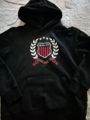 Svart hoodie från GANT, stl 158/164 - Svart hoodie från GANT med stort broderat emblem på bröstet, vita och röda detaljer samt texten 'Established 1949'. Klassisk känguruficka framtill och huva. Perfekt för en sportig och avslappnad stil.