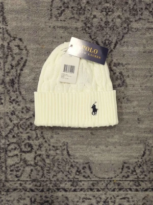 Vit kabelstickad mössa Polo Ralph Lauren - Snygg vit kabelstickad mössa från Polo Ralph Lauren med uppvikt kant och klassisk broderad logga i marinblått framtill. Tillverkad i mjukt material som håller dig varm och stilren under kalla dagar.
