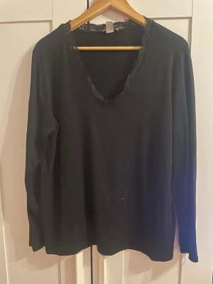 Svart långärmad tröja med spets - En svart långärmad tröja med v-ringning och snygg spetsdetalj runt halsen. Tröjan har en enkel och stilren design som passar till det mesta. Materialet ser mjukt och bekvämt ut, perfekt för lager på lager under kallare dagar.