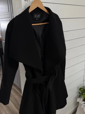 Svart kappa  med skärp - Stilren svart kappa med bred krage och knytband i midjan. Kappan har slät insida med foder och en klassisk, elegant siluett. Perfekt för dig som vill ha en tidlös och trendig look.