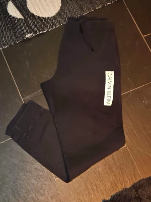 Svarta mjukisbyxor Calvin Klein - Säljer ett par svarta mjukisbyxor från Calvin Klein med resår i midjan och dragsko. Byxorna har en vit logga på vänster ben och muddar vid bensluten. Perfekta för chill dagar och riktigt skön passform.