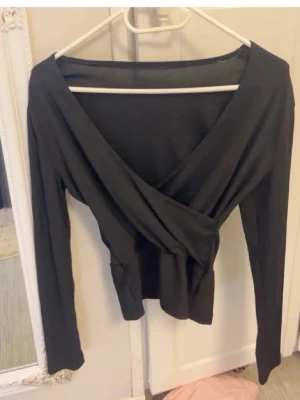 Svart omlottopp med v-ringning - Snygg svart omlottopp med djup v-ringning och långa ärmar. Toppen har en croppad passform och är tillverkad i ett mjukt, stretchigt material som sitter tight mot kroppen. Perfekt för att skapa en trendig och stilren look.